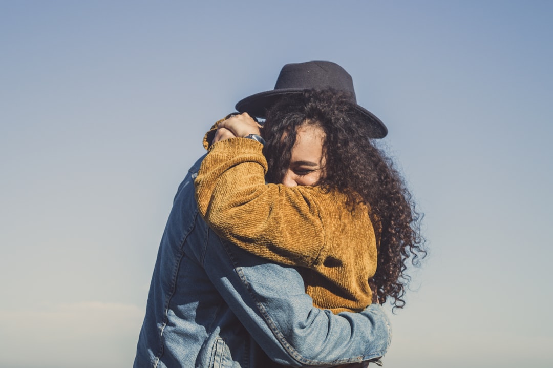 Adult"A close-up outdoor shot of a person with long curly hair and a mustard yellow corduroy sweater hugging another person wearing a black hat and a blue denim jacket. The two are embracing tightly against a clear, light blue sky background."