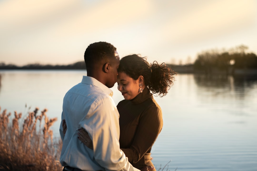 "Couple embracing affectionately near a calm lake at sunset, with warm golden light reflecting on the water."