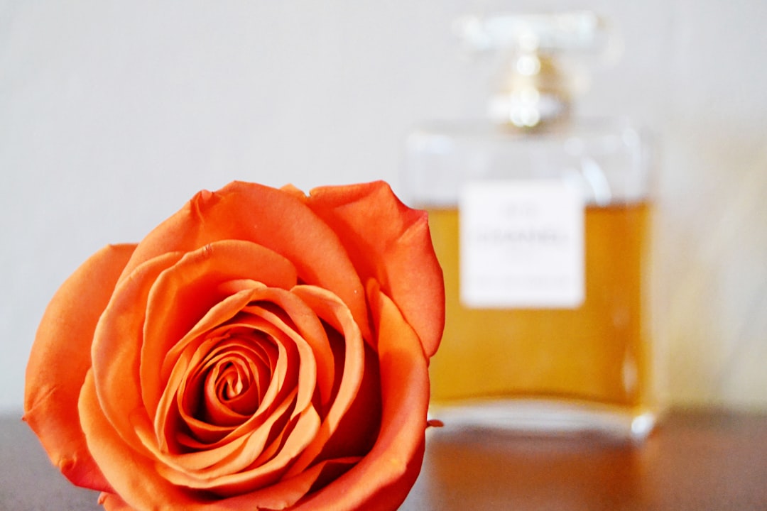 A close-up, low-angle shot of a vibrant orange rose in the foreground, with a clear glass perfume bottle with a golden liquid and light cap blurred in the background.