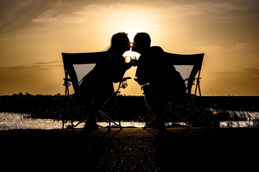 Silhouette of a couple sitting on camping chairs, sharing a drink while watching the sunset by the water.