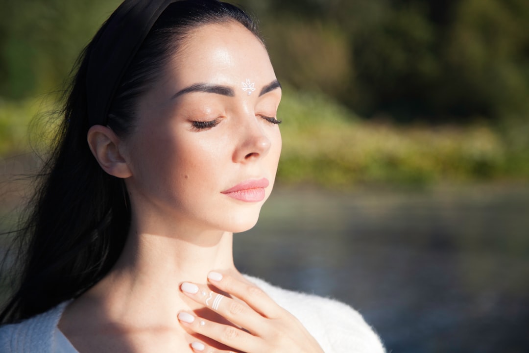 A close-up of a woman with her eyes closed in peaceful contemplation, wearing a dark headband and white top, with a small white design on her forehead and hand, against a blurred natural background.