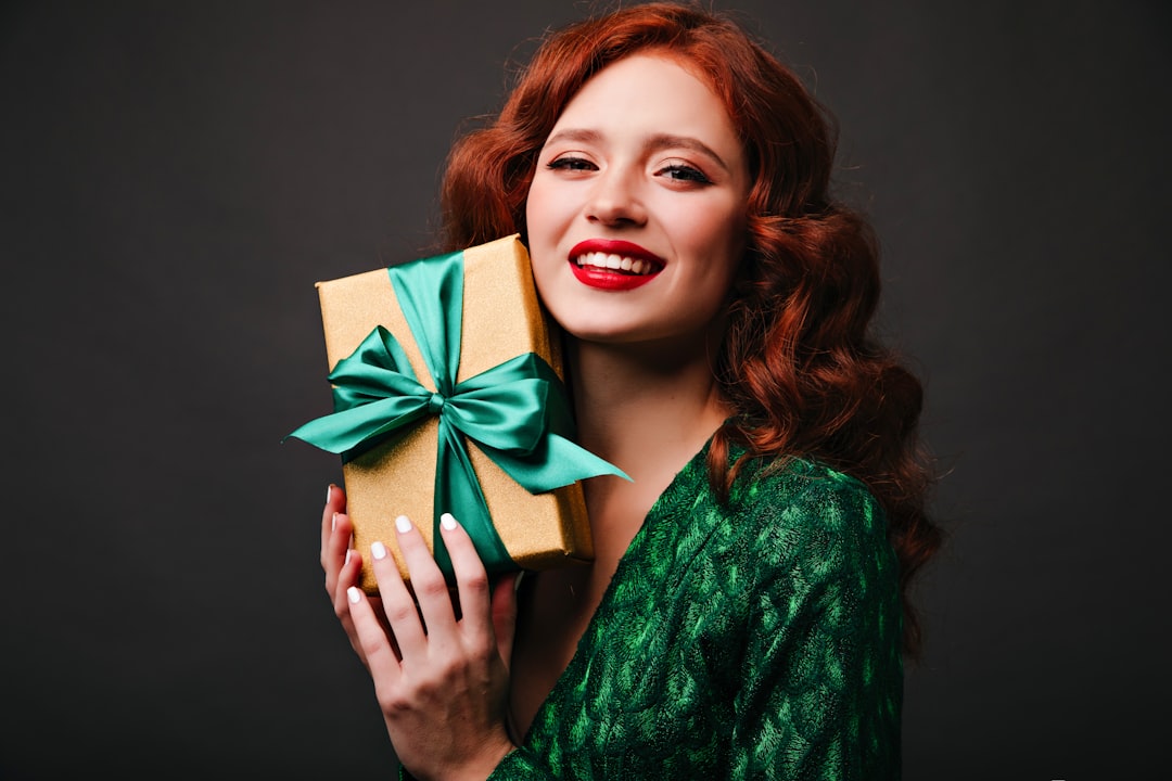 A vibrant, smiling woman with long red hair, wearing a sparkly green garment, holds a gold-wrapped gift box tied with a satin green ribbon close to her cheek. She has bright red lipstick and white nail polish, set against a dark grey background.