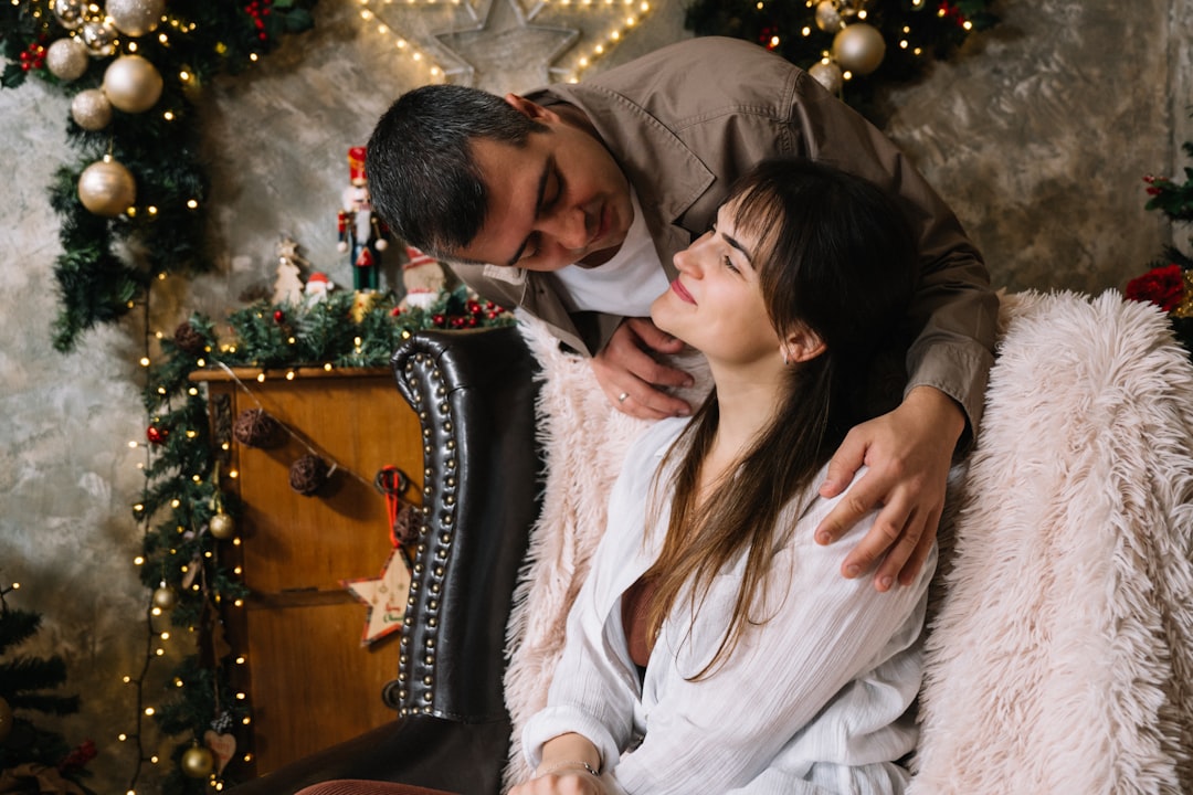 She is seated in a large, plush armchair (partially visible, dark leather/fabric) and wears a light-colored, possibly white, open shirt. She has dark hair with bangs and is smiling gently with her eyes closed, resting her head against the man's shoulder.
