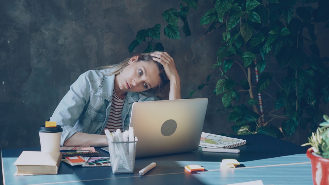 A tired young woman sitting at a desk with her laptop, resting her head on her hand, surrounded by notebooks, stationery, and a coffee cup, appearing exhausted or stressed while working.